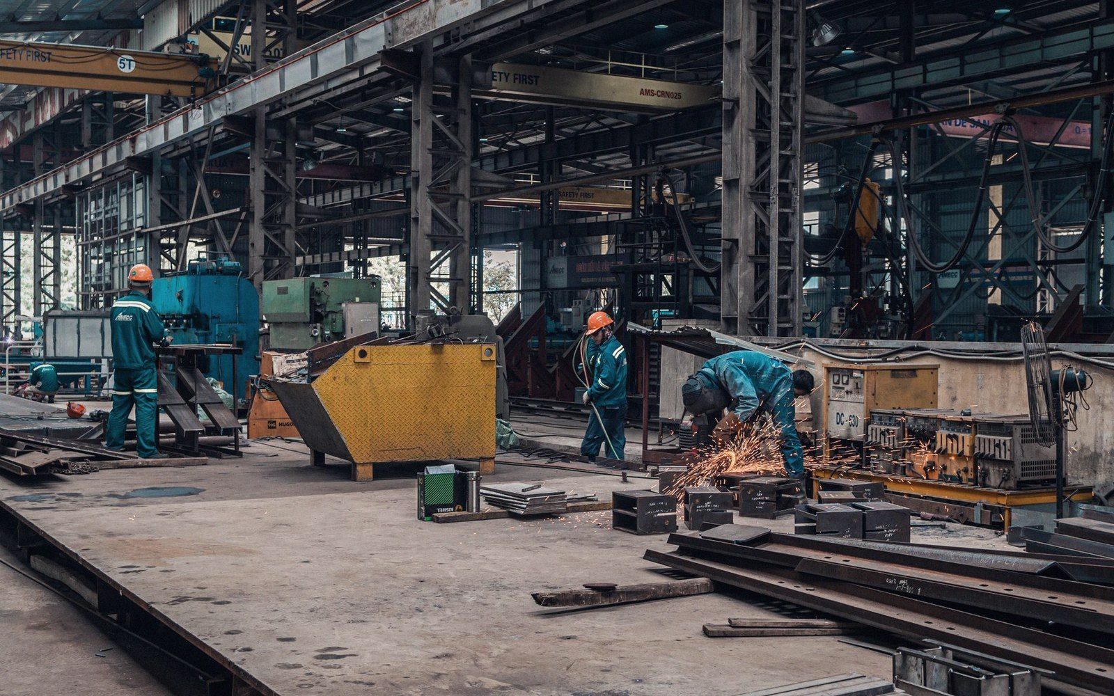 Factory floor with metal fabrication machinery, adult workers in overalls, industrial strip lighting from above