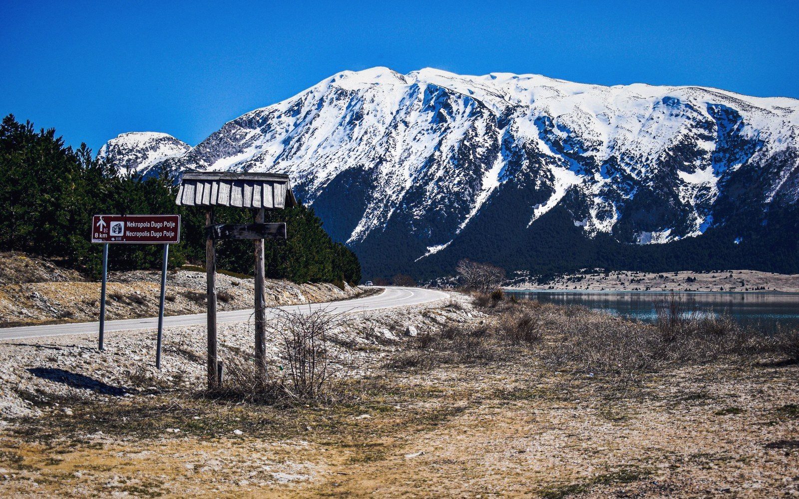 Road signs pointing to Dugo Polje necropolis along a lakeside road below snow-capped mountains