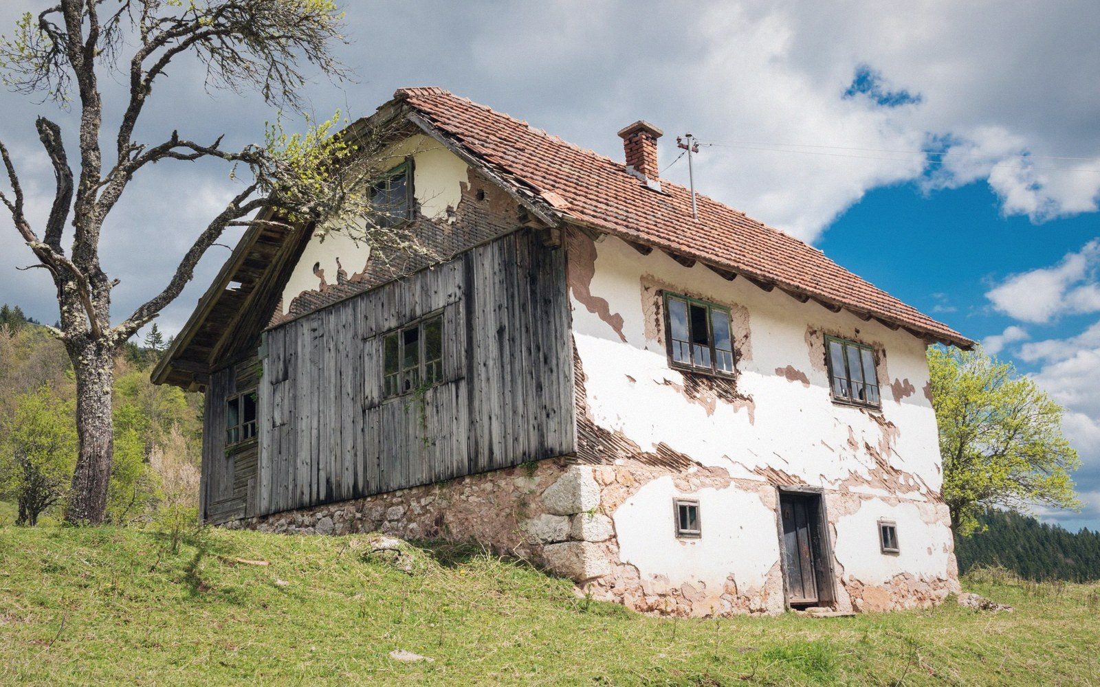 Weathered farmhouse with peeling plaster and wooden gable stands on a grassy hillside