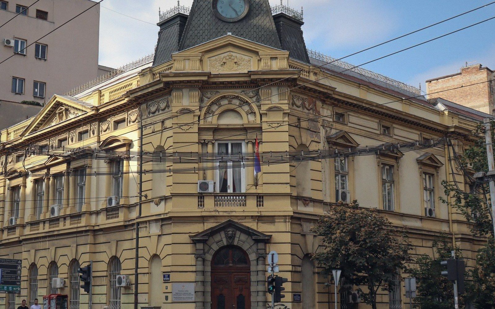 Ornate yellow neoclassical building with clock tower and Serbian flag on corner facade