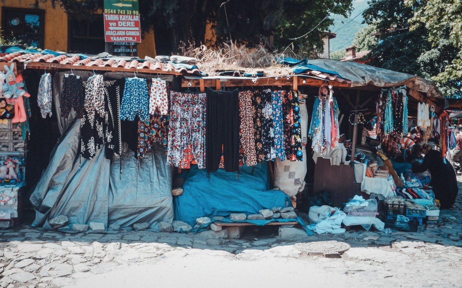 Patterned scarves and fabrics hang from a roadside market stall under a tiled roof