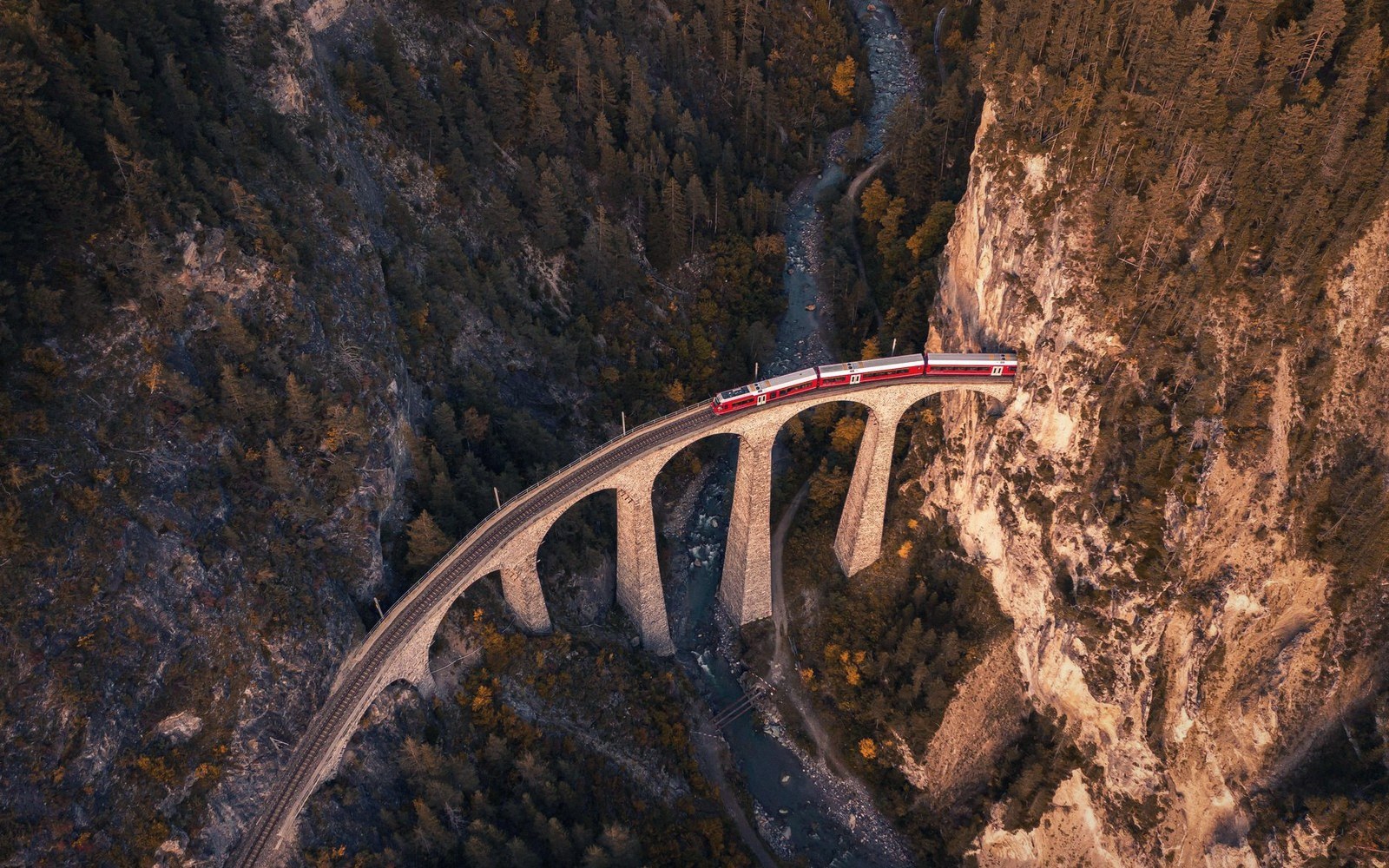 A red train crosses a stone viaduct over a forested mountain gorge