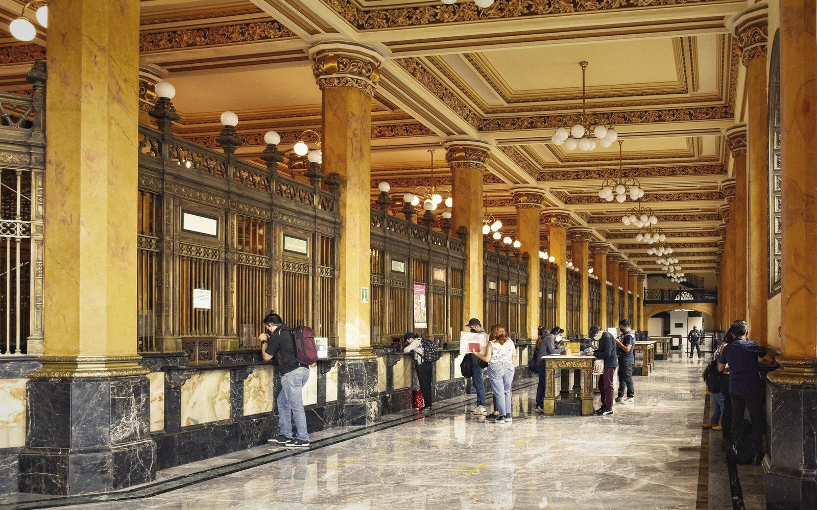 People wait at ornate teller counters in a marble-columned post office hall