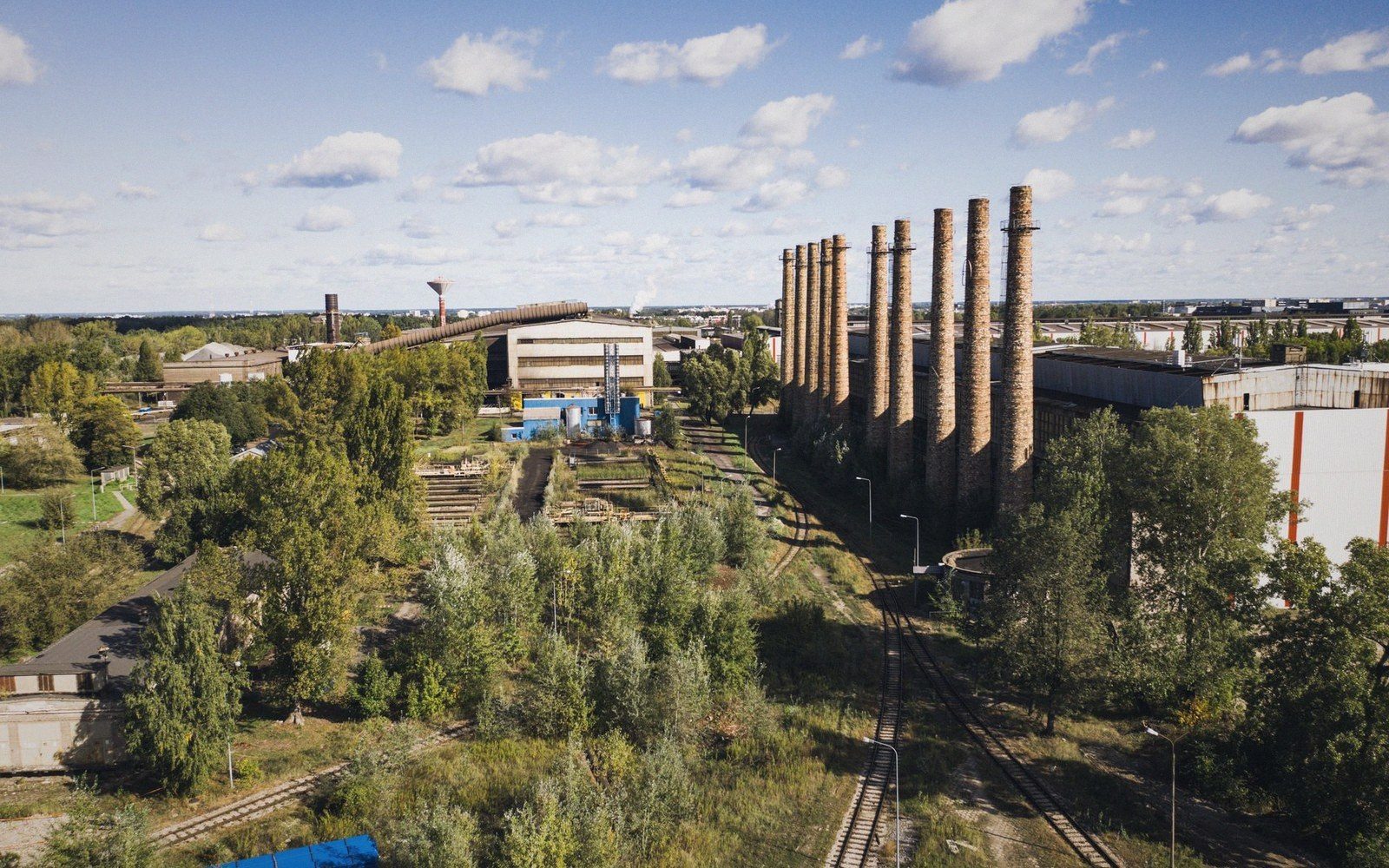 Row of tall brick chimneys beside abandoned industrial buildings and overgrown rail tracks