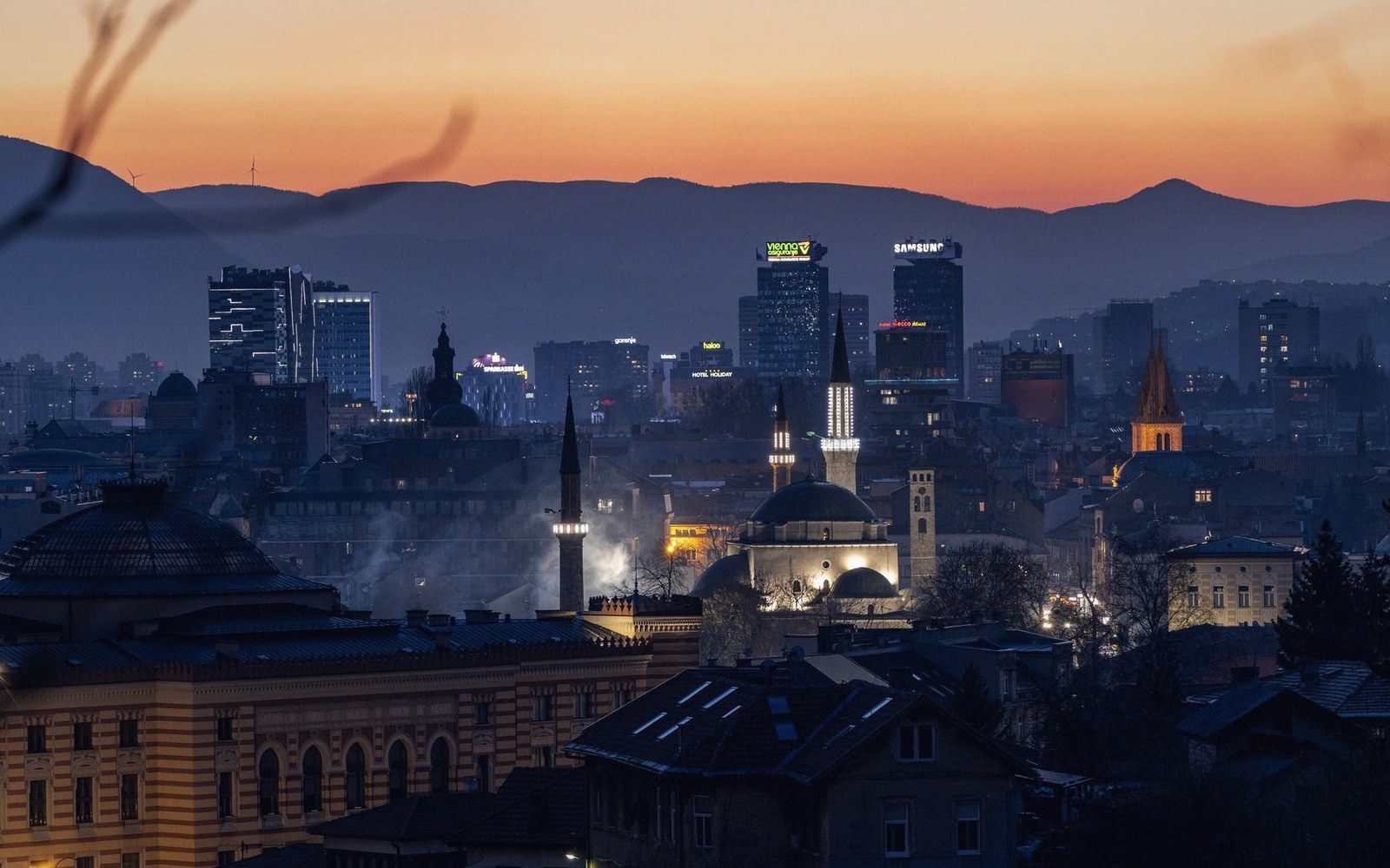 City skyline at dusk with a mosque minaret and illuminated skyscrapers against mountains