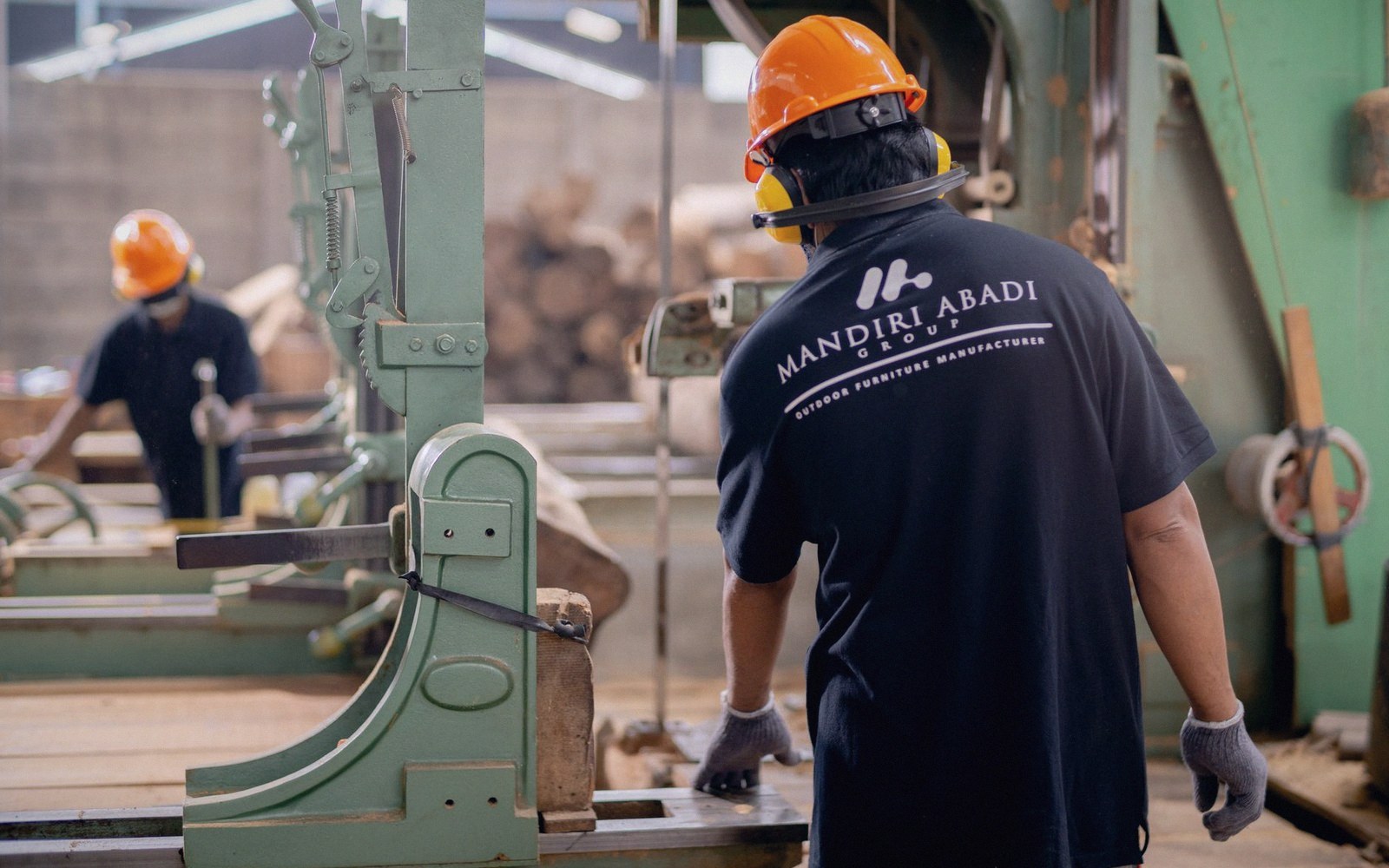 Workers in hard hats and ear protection operate a bandsaw at a Mandiri Abadi Group sawmill
