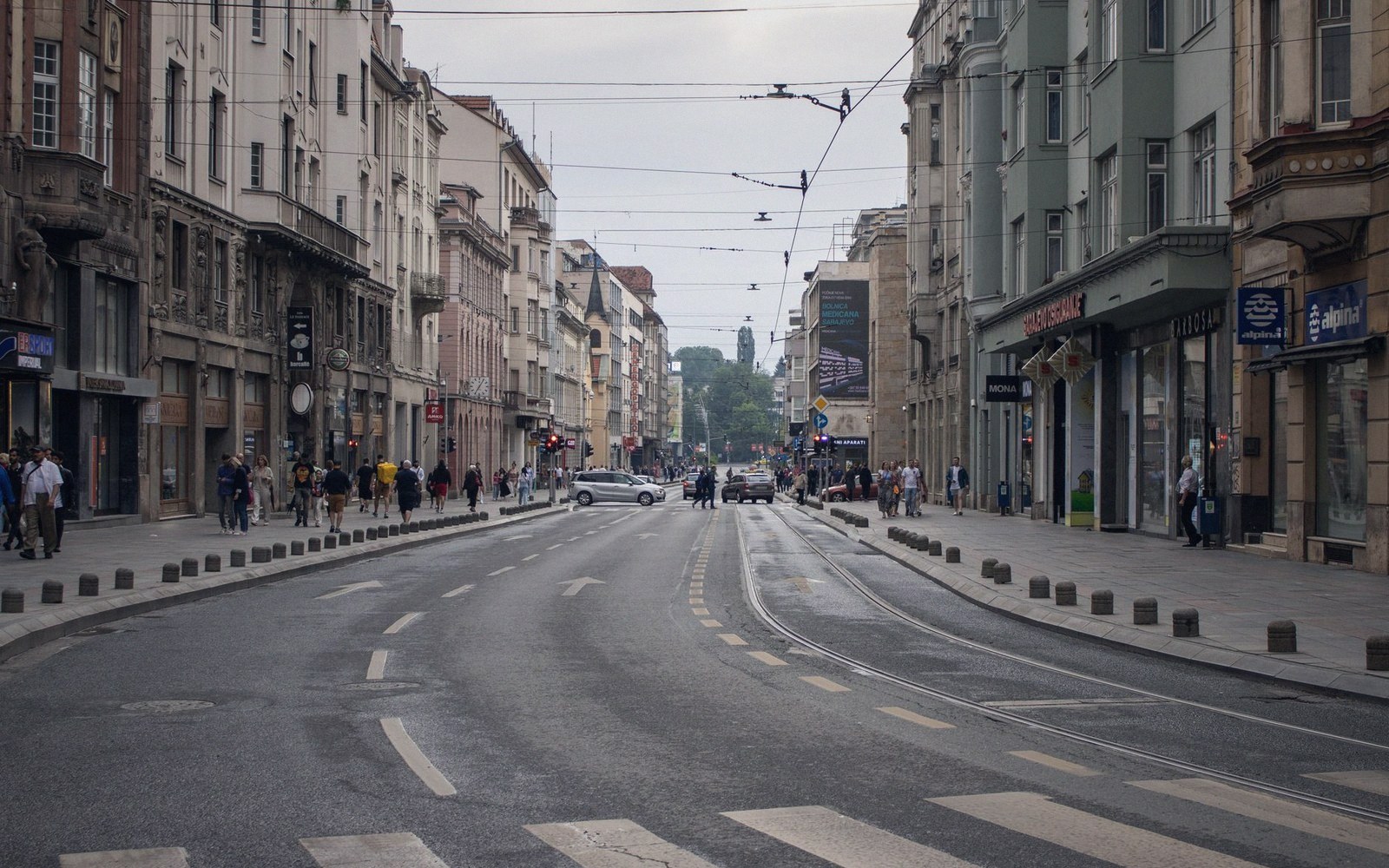 Pedestrians cross a wide city street lined with shops and tram wires overhead