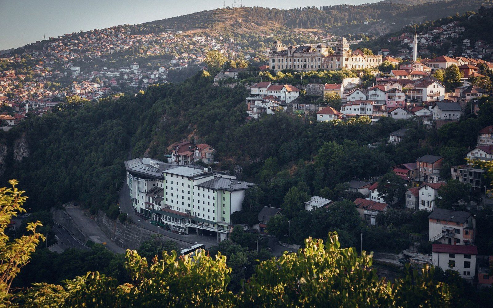 Sarajevo seen from a hillside at late afternoon, apartment blocks descending into a valley, grey-green hills behind